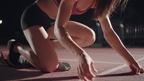 La-Mujer-Al-Inicio-De-La-Carrera-Se-Pone-Las-Almohadillas,-Se-Levanta-Y-Corre-A-Cámara-Lenta-Por-La-Noche-En-El-Estadio.-La-Corredora-Se-Agacha-En-La-Posición-Inicial-Antes-De-Comenzar-A-Correr.